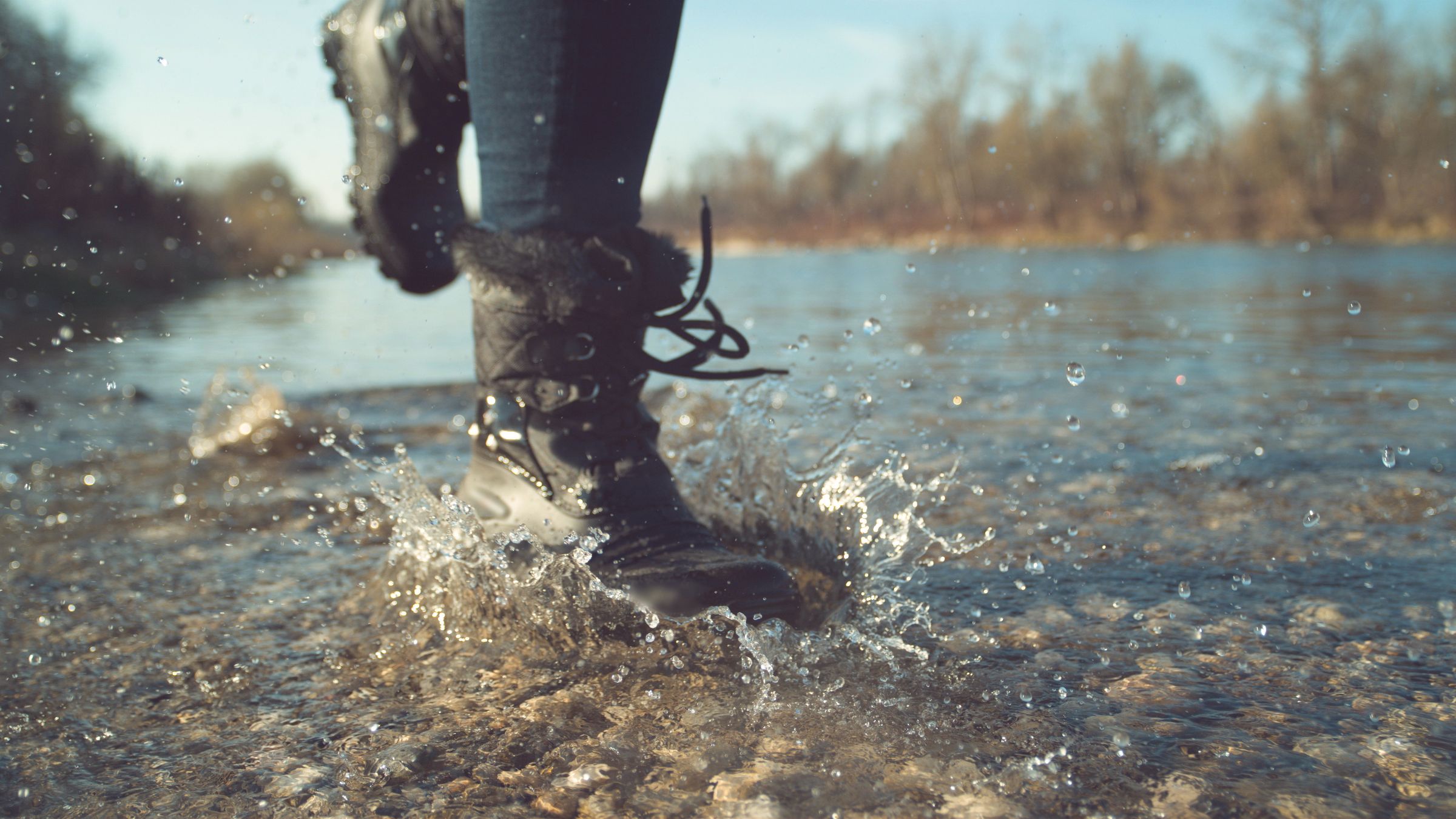 Close-up of a person's legs in black fur-lined boots splashing in a shallow, rocky river.