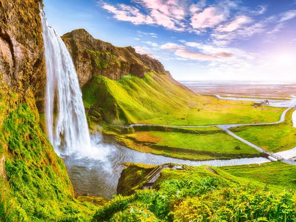 Scenic view of a tall waterfall cascading down a green cliff into a river that flows through a lush valley under a colorful sky.
