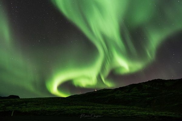 Green aurora borealis swirls across a dark, starry sky above a dark landscape.