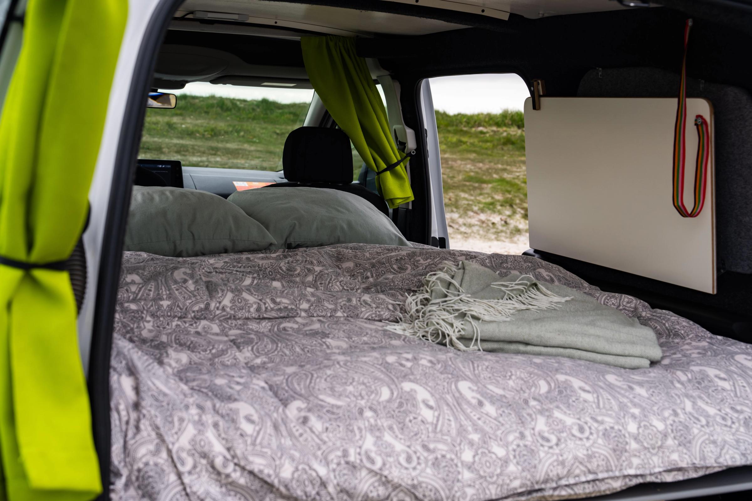 Bett mit gemusterter Bettdecke, Kissen und einer zusammengelegten Tagesdecke, daneben ein verstautes Holzbrett