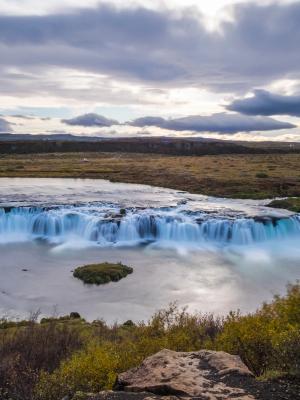 A wide waterfall with milky water cascades through a sparse, autumnal landscape under a cloudy sky.