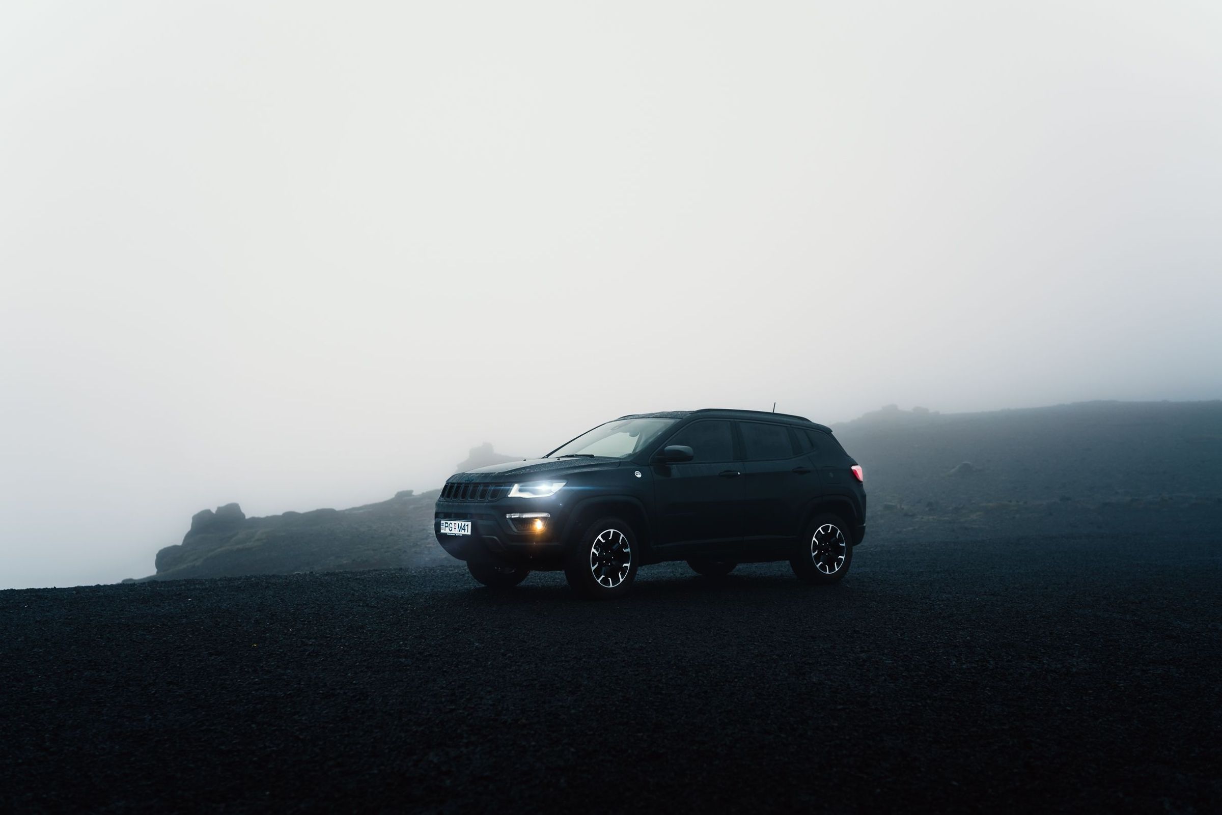 A dark Jeep Compass SUV with headlights on, parked on a dark, foggy landscape.