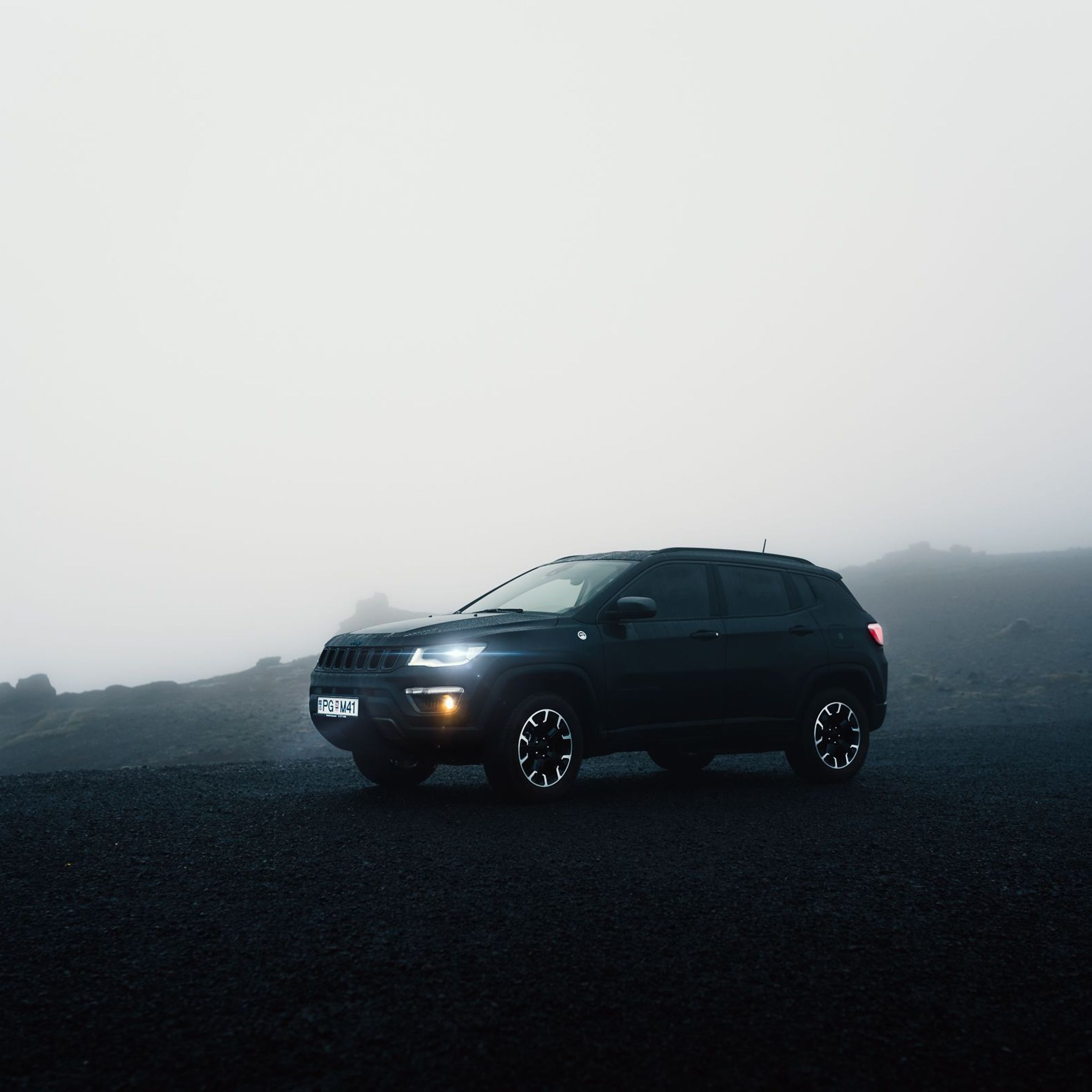 A dark Jeep Compass SUV with headlights on, parked on a dark, foggy landscape.
