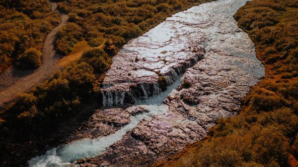 Aerial view of a river with multiple waterfalls flowing through reddish-brown foliage, with a winding dirt path on the left.