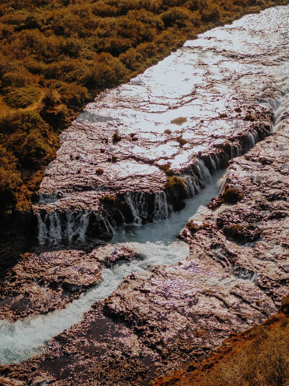 Aerial view of a river with multiple waterfalls flowing through reddish-brown foliage, with a winding dirt path on the left.
