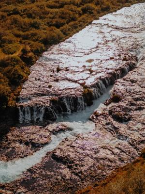 Aerial view of a river with multiple waterfalls flowing through reddish-brown foliage, with a winding dirt path on the left.