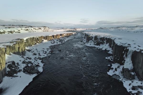 Un poderoso río atraviesa un cañón cubierto de nieve, rodeado de acantilados rocosos y con una cascada a lo lejos.