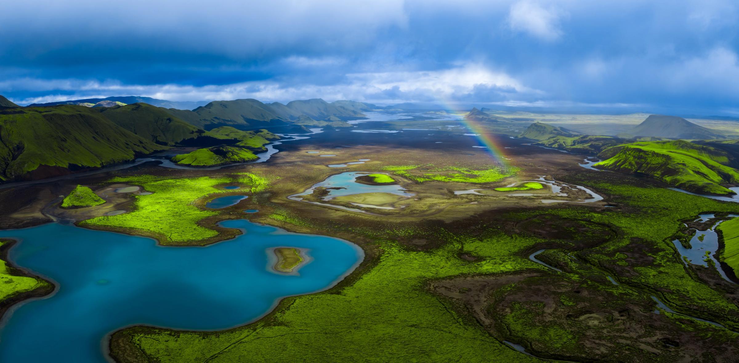 Aerial view of a vibrant green landscape with blue lakes, rivers, mountains, and a rainbow under a cloudy sky.
