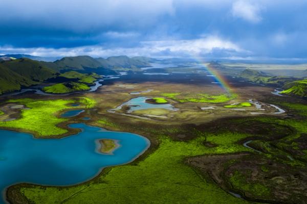 Aerial view of a vibrant green landscape with blue lakes, rivers, mountains, and a rainbow under a cloudy sky.