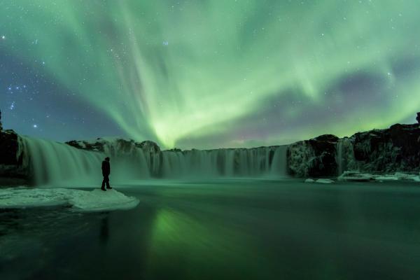 a man is standing in front of a waterfall under the aurora borealis .