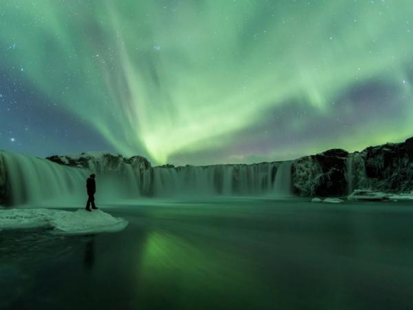 Green Northern Lights illuminate a wide waterfall and a person standing on an ice floe.