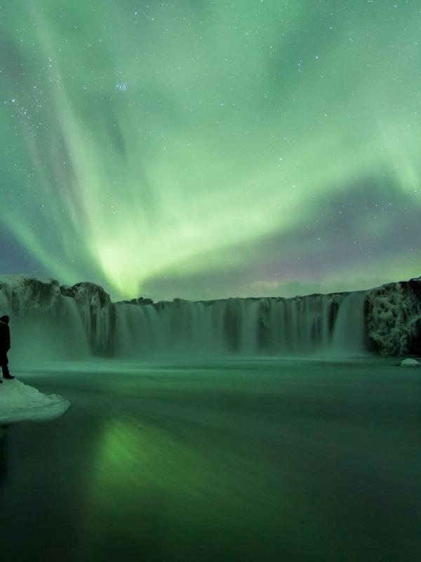 un hombre está parado frente a una cascada bajo la aurora boreal.