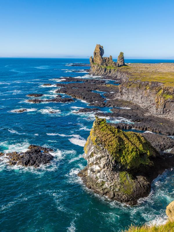Dramatic coastal landscape with dark jagged rocks, towering sea stacks, and cliffs along a deep blue ocean.