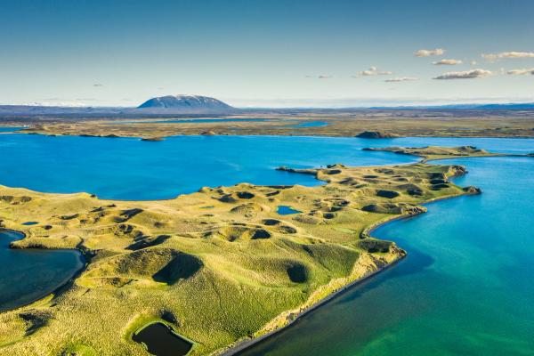 Aerial view of a bright blue lake, green landscape with many small crater-like mounds, and a distant snow-capped mountain.