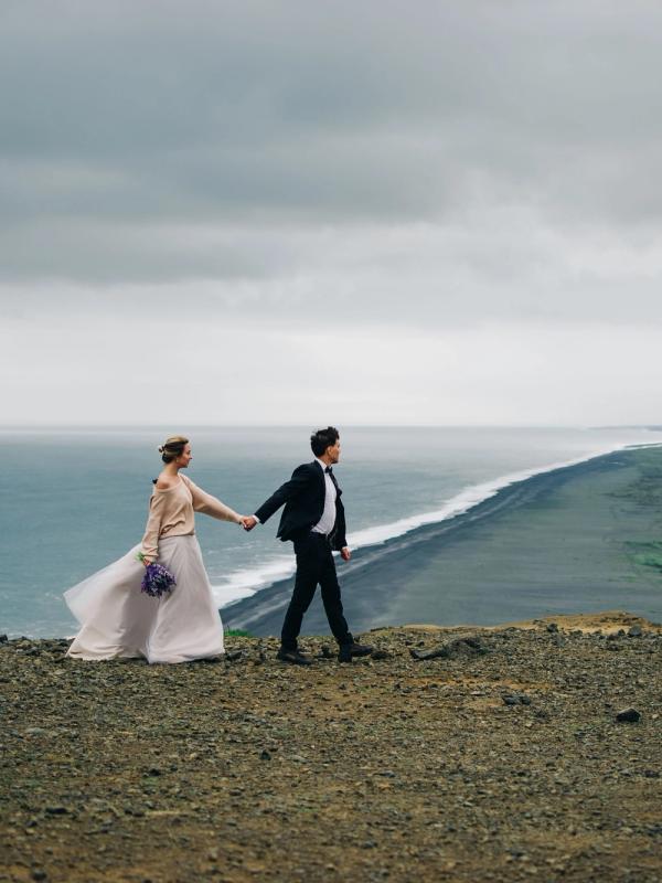 a bride and groom are walking on top of a hill overlooking the ocean .