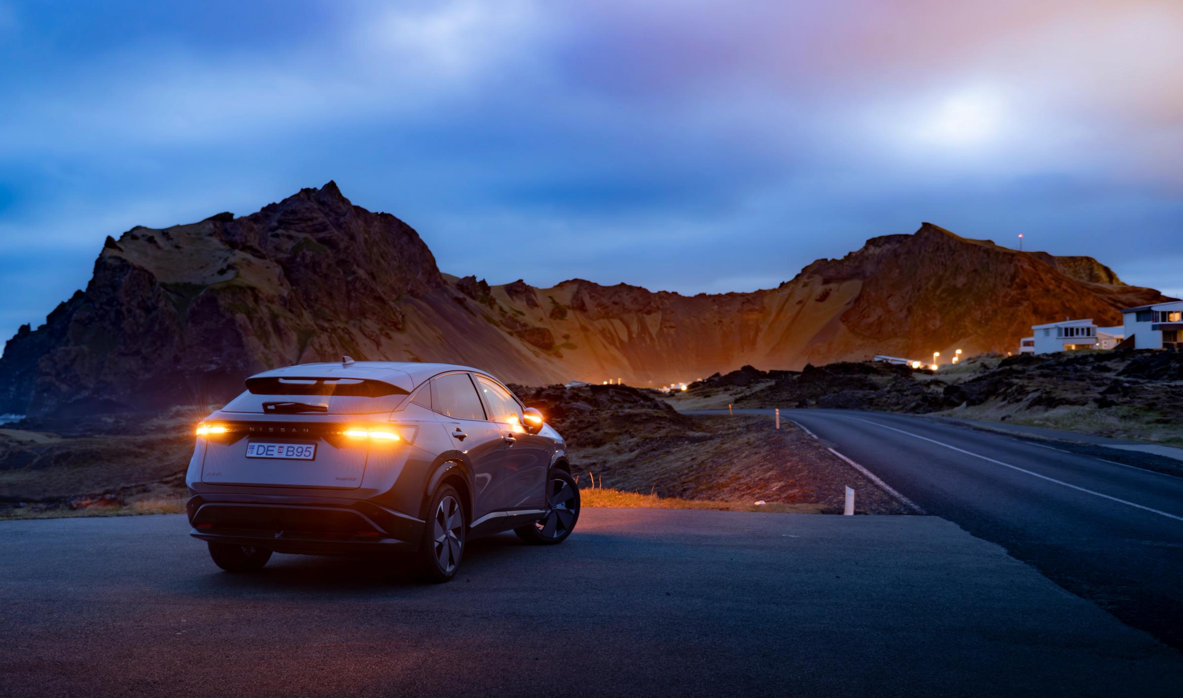 Nissan Ariya séður aftan frá, lagt við malbikaðan veg með upplýst fjöll í bakgrunni í ljósaskiptunum