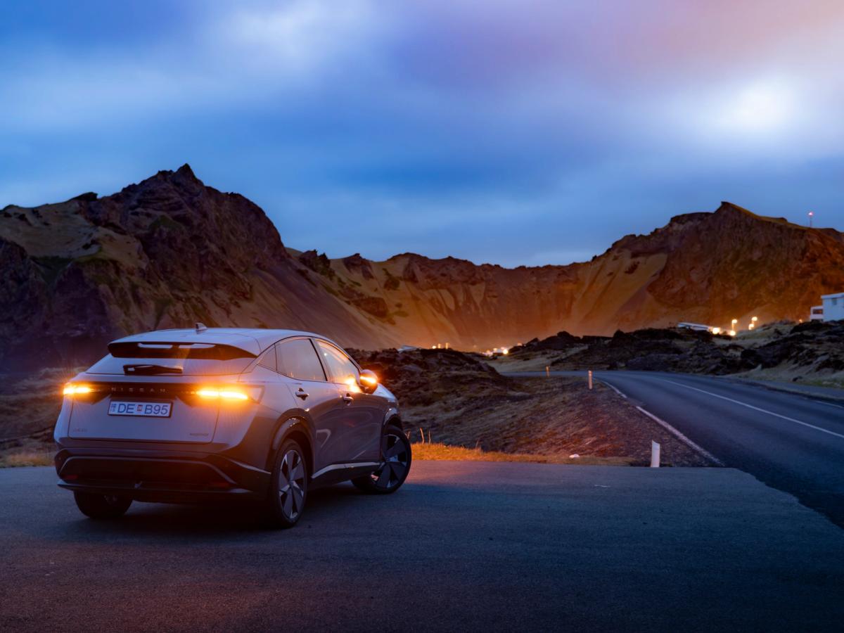 Nissan Ariya á fjallvegi að kvöldi Nissan Ariya séður aftan frá, lagt við malbikaðan veg með upplýst fjöll í bakgrunni í ljósaskiptunum
