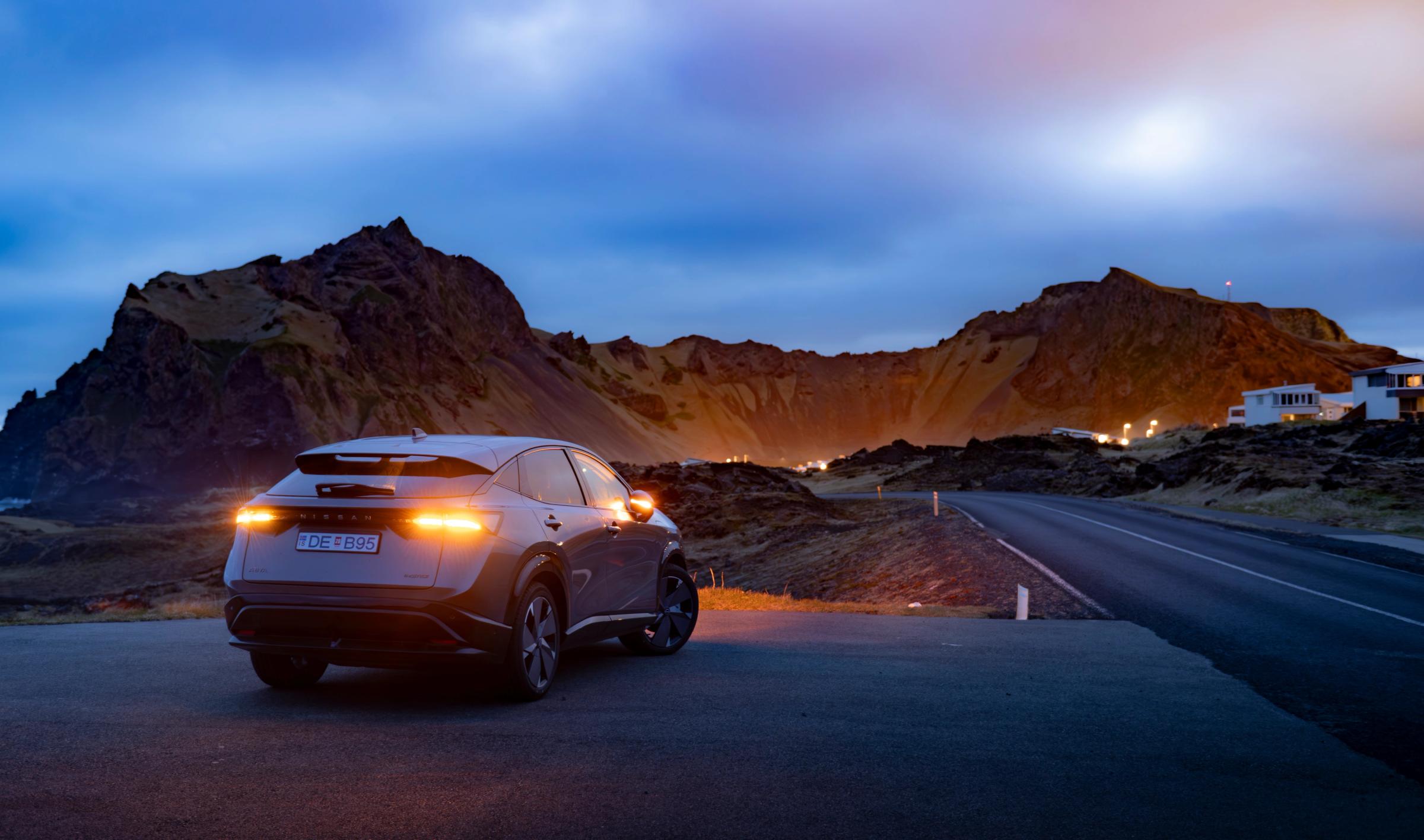 Nissan Ariya á fjallvegi að kvöldi Nissan Ariya séður aftan frá, lagt við malbikaðan veg með upplýst fjöll í bakgrunni í ljósaskiptunum