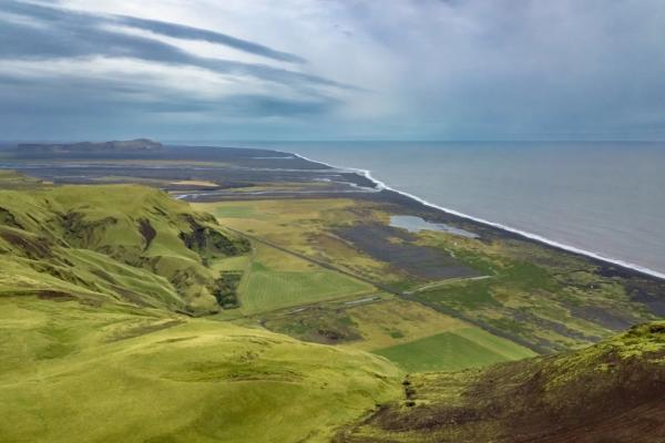 Vast landscape with green hills descending to a dark coastal plain, black sand beach, and ocean under a cloudy sky.