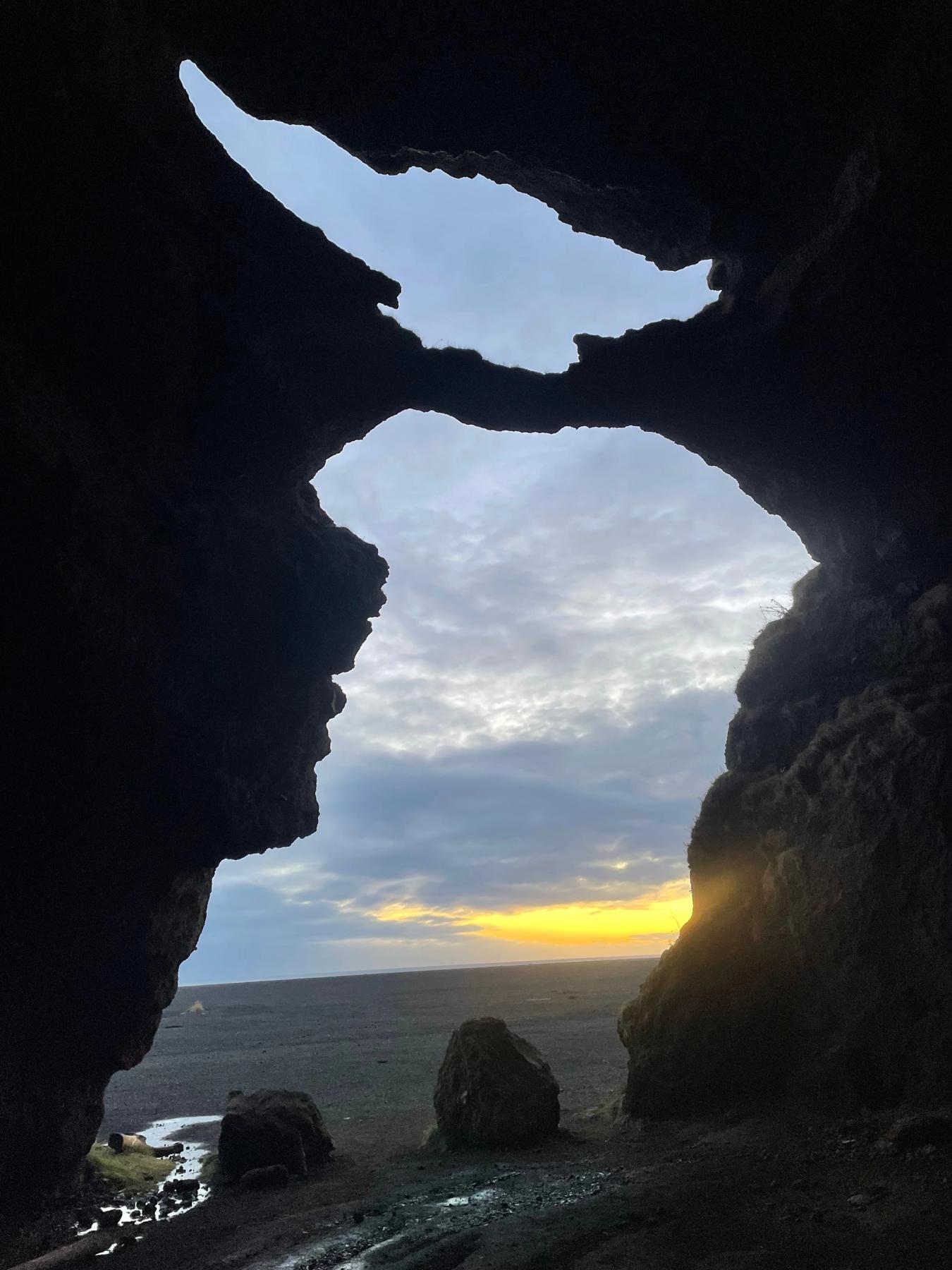 View from inside a dark cave, looking out at a flat, dark landscape under a cloudy sky with a yellow sunset on the horizon.