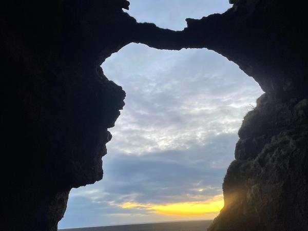 View from inside a dark cave, looking out at a flat, dark landscape under a cloudy sky with a yellow sunset on the horizon.