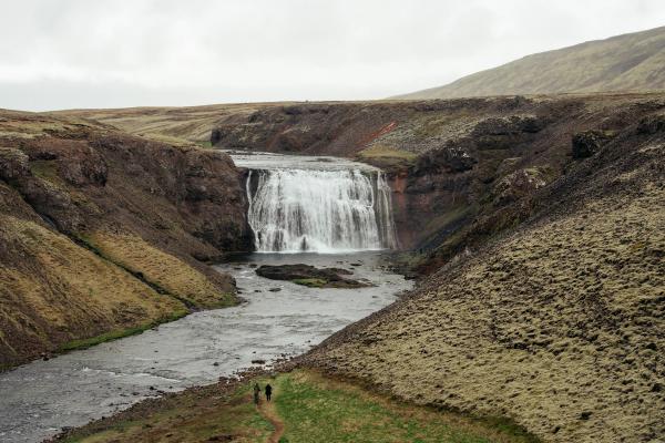 A wide waterfall plunges into a river flowing through a rugged valley, with two small hikers on a path.