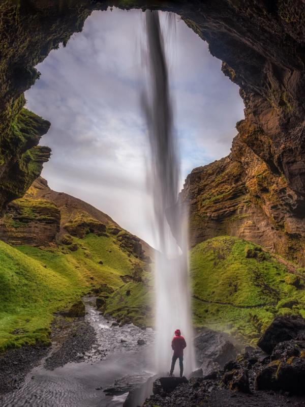 A person in a red jacket stands under a tall waterfall, viewed from inside a dark cave with green hills beyond.