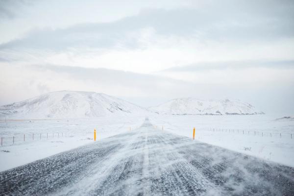 Una carretera cubierta de nieve con ventisca conduce a montañas distantes coronadas de nieve bajo un cielo nublado.