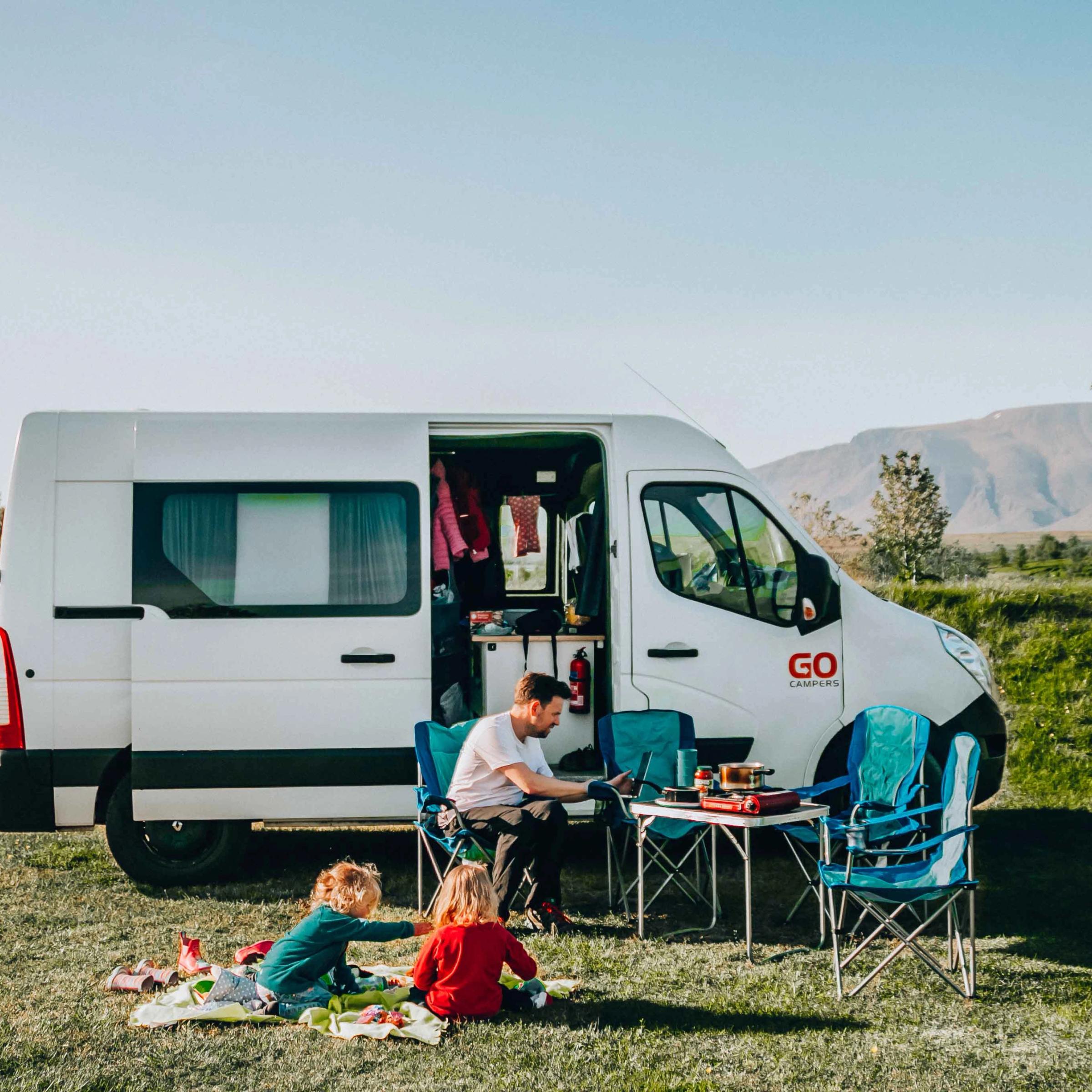 A man works on a laptop at a camping table next to a white campervan while two children play on a blanket on the grass.