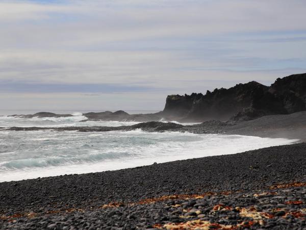 A black pebble beach with white waves crashing against dark rocky cliffs under a cloudy sky.
