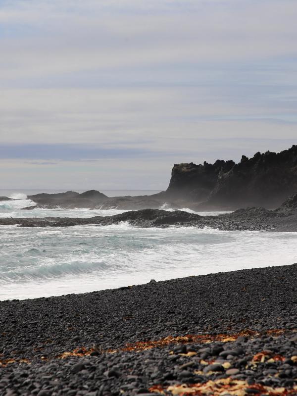 A black pebble beach with white waves crashing against dark rocky cliffs under a cloudy sky.