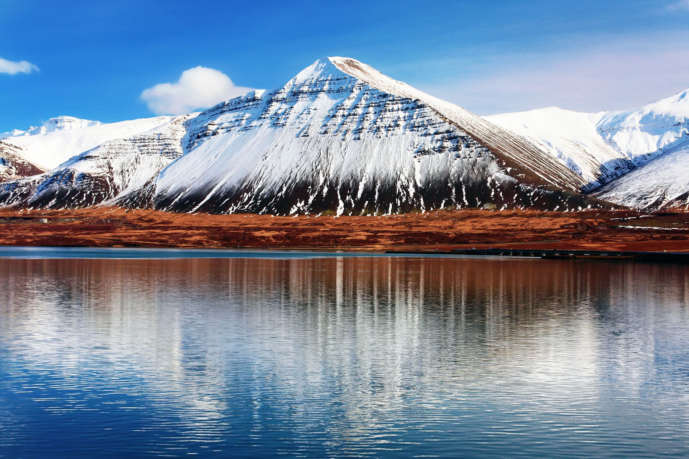 Montañas nevadas con colinas rojizas reflejadas en aguas tranquilas bajo un cielo azul.