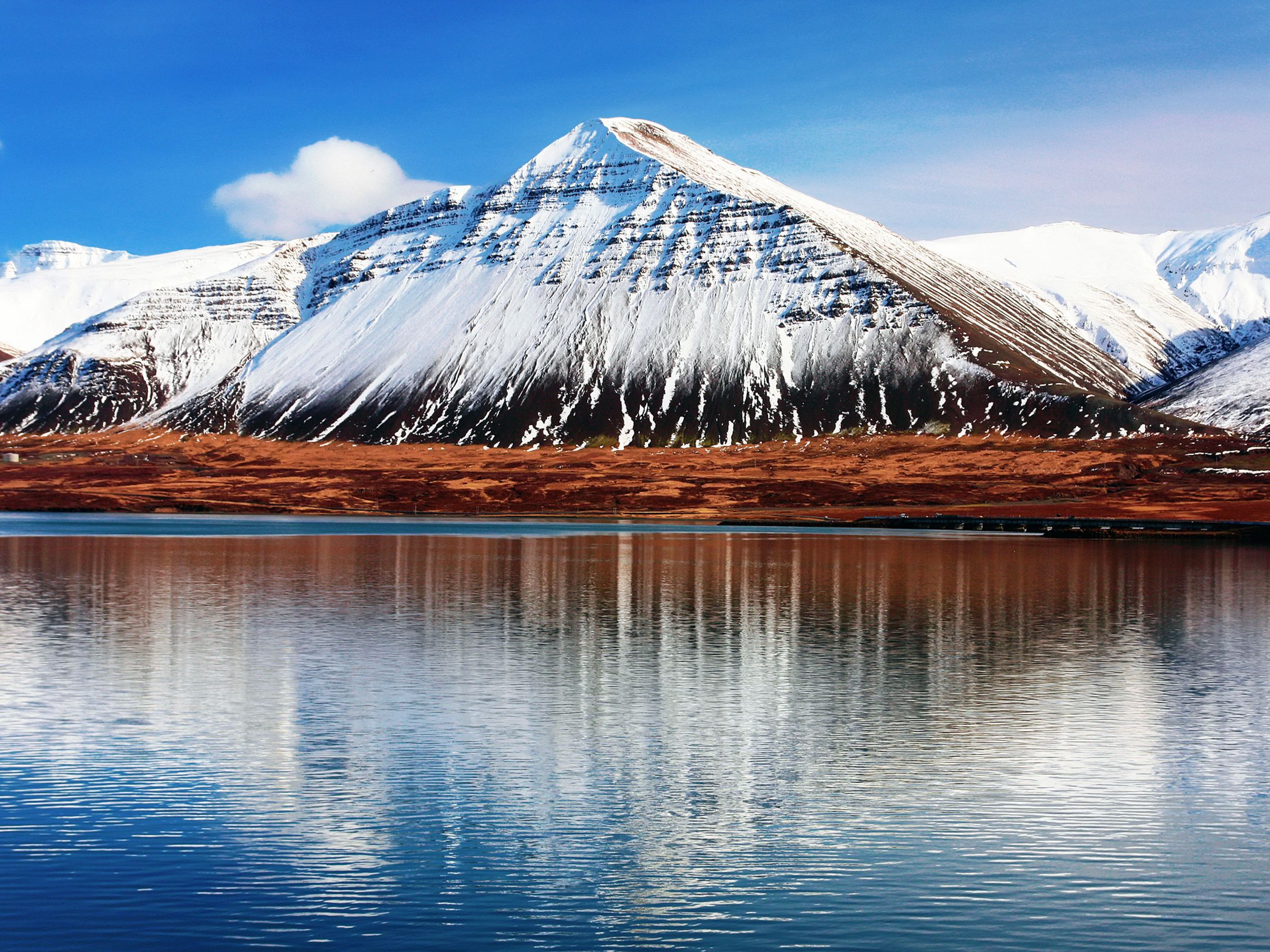 Montañas nevadas con colinas rojizas reflejadas en aguas tranquilas bajo un cielo azul.