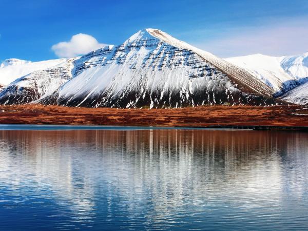 Snow-capped mountains with reddish-brown foothills reflected in calm water under a blue sky.