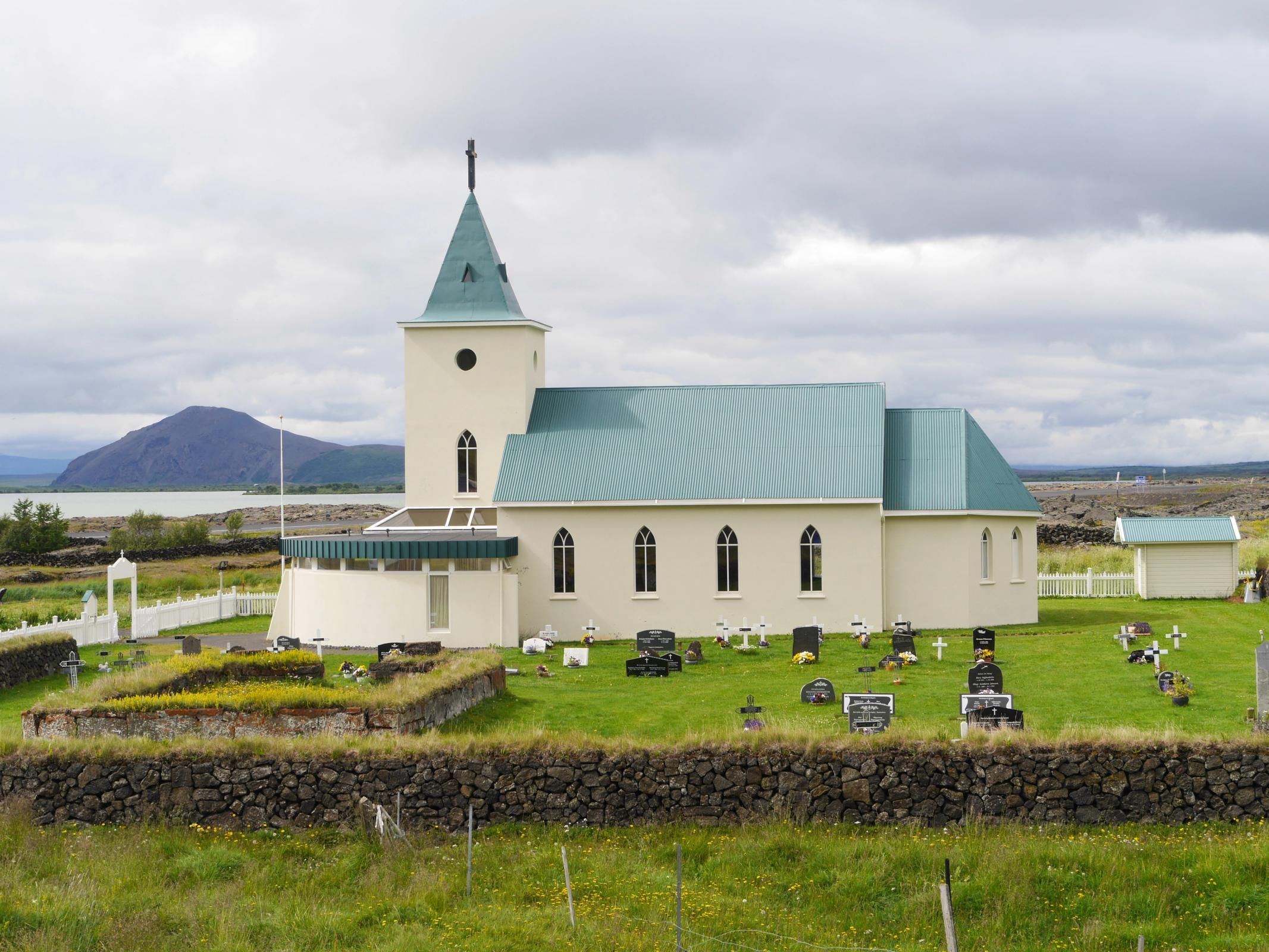 Una iglesia blanca con techo y campanario verdes se alza en un cementerio, con un lago y una montaña de fondo bajo un cielo nublado.