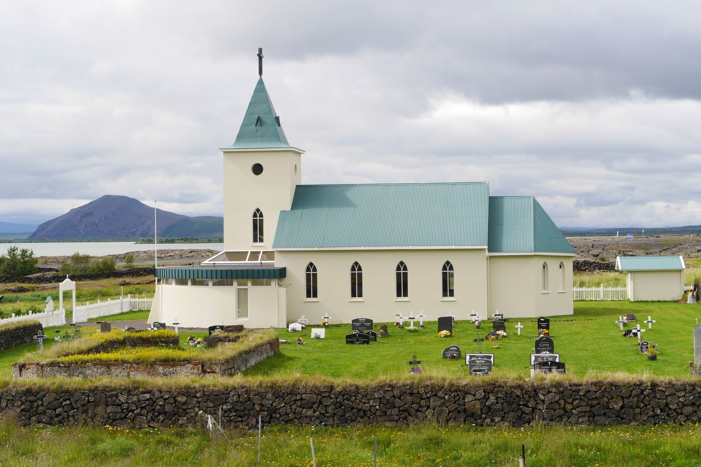 Una iglesia blanca con techo y campanario verdes se alza en un cementerio, con un lago y una montaña de fondo bajo un cielo nublado.