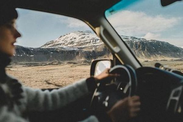 a woman is driving a car on a dirt road with mountains in the background .