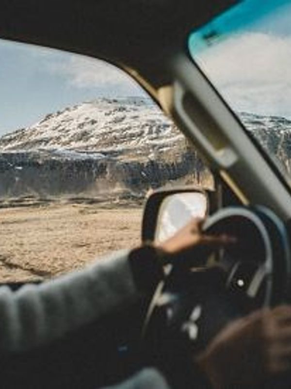 A person driving a car looks out at snow-capped mountains and a vast, barren landscape.