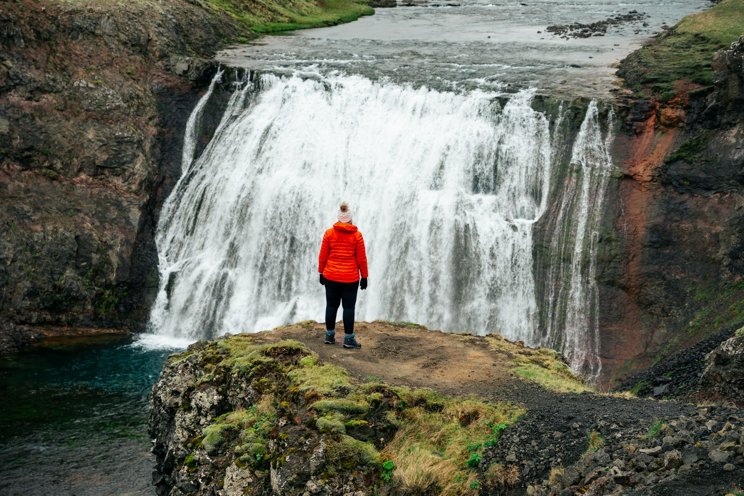 A person in an orange jacket stands on a cliff overlooking a powerful waterfall.