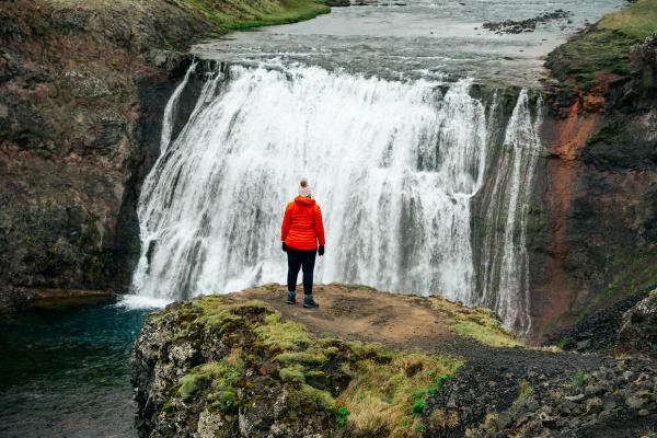 A person in an orange jacket stands on a cliff overlooking a powerful waterfall.