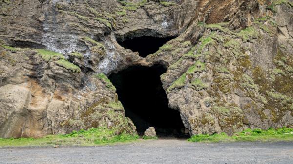 Two dark cave entrances in a rugged, moss-covered rock face above a gravel path.