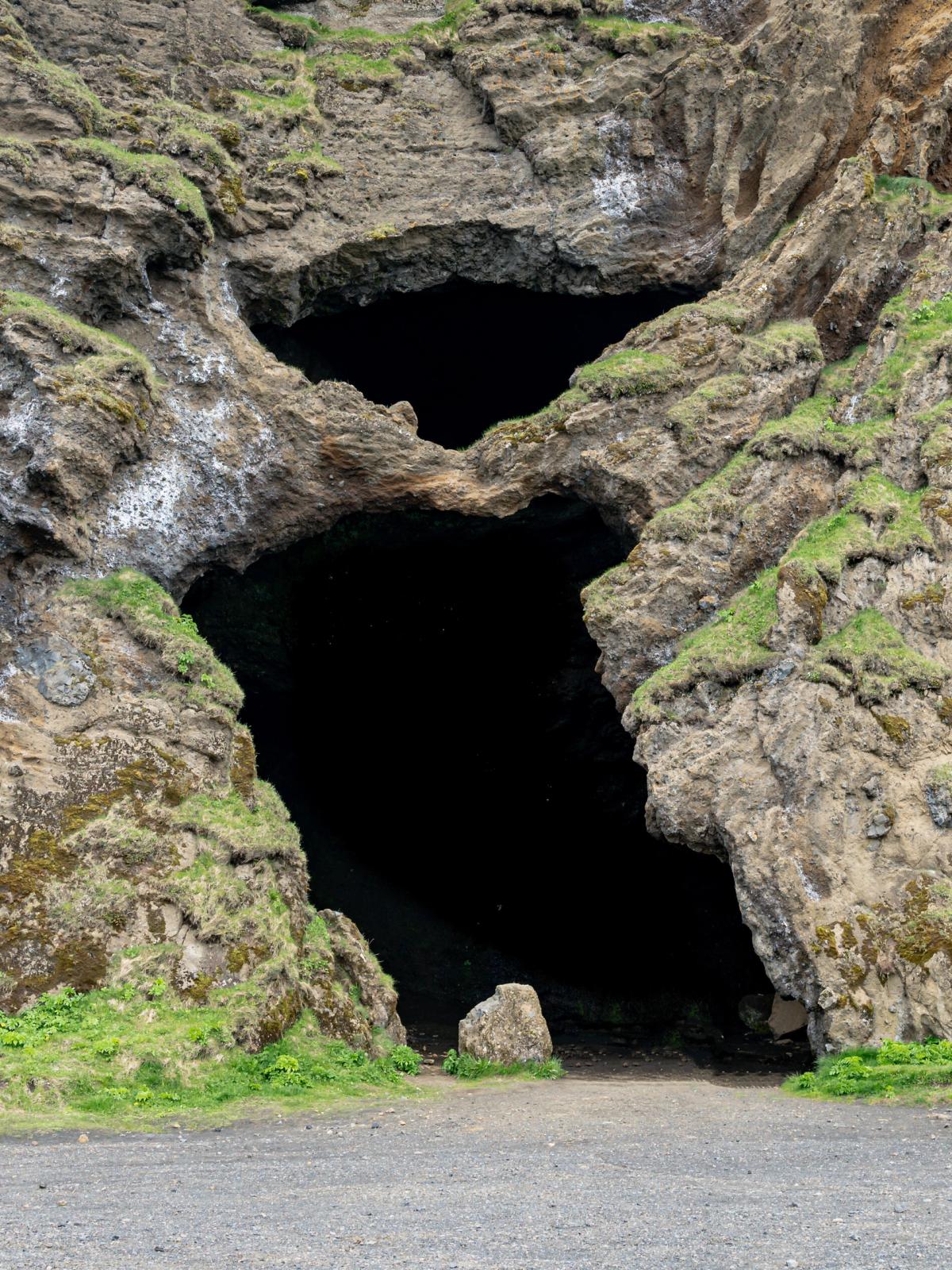 Two dark cave entrances in a rugged, moss-covered rock face above a gravel path.