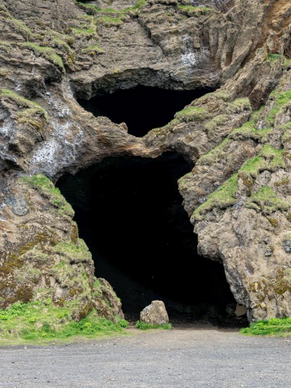Two dark cave entrances in a rugged, moss-covered rock face above a gravel path.