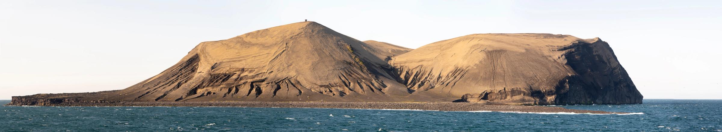 A barren island with sandy slopes and dark, eroded cliffs, surrounded by blue ocean under a pale sky.
