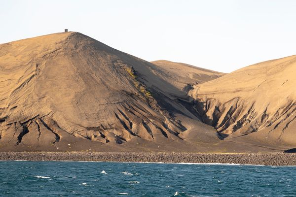 A barren island with sandy slopes and dark, eroded cliffs, surrounded by blue ocean under a pale sky.