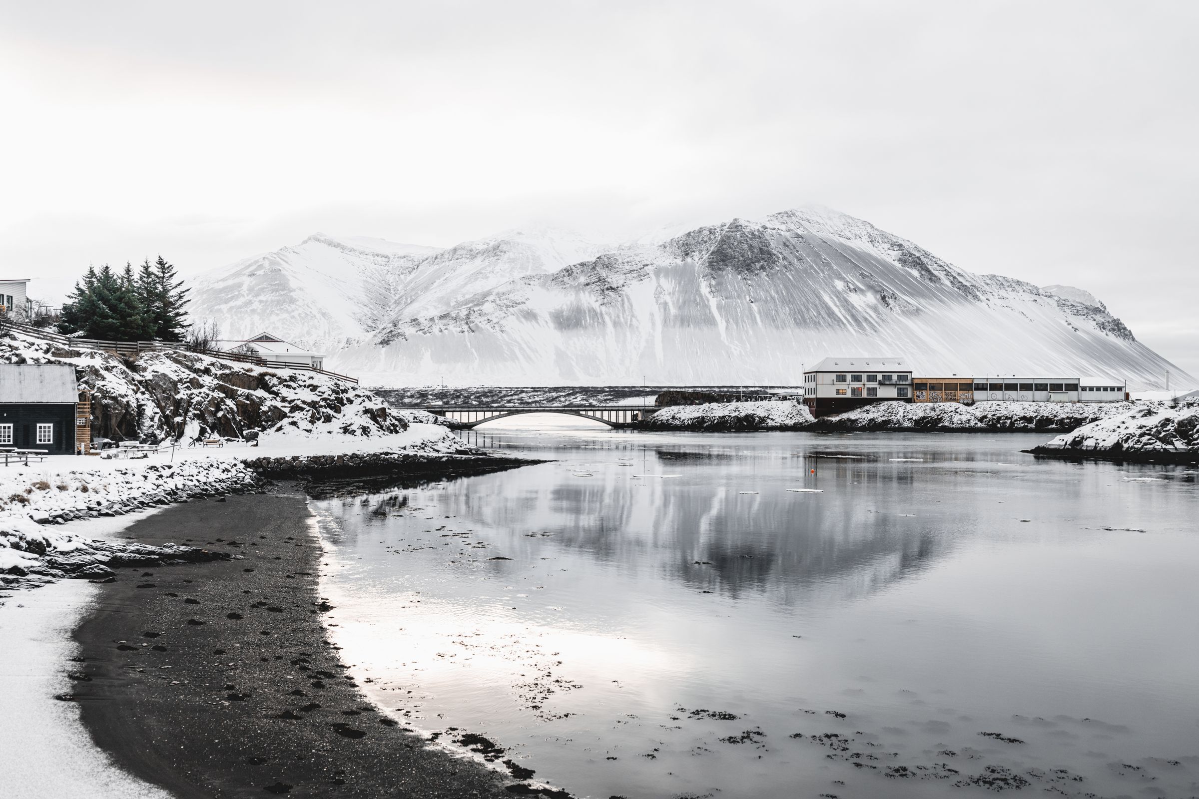 Montañas nevadas y un pueblo costero reflejados en aguas tranquilas, cruzados por un puente.