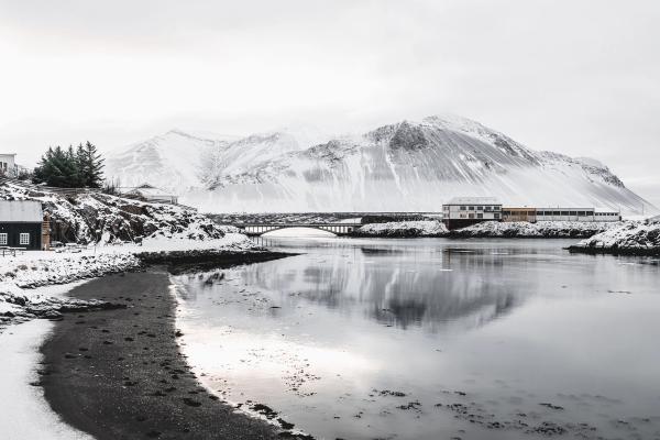 Snowy mountains and a coastal village reflected in calm water, crossed by a bridge.