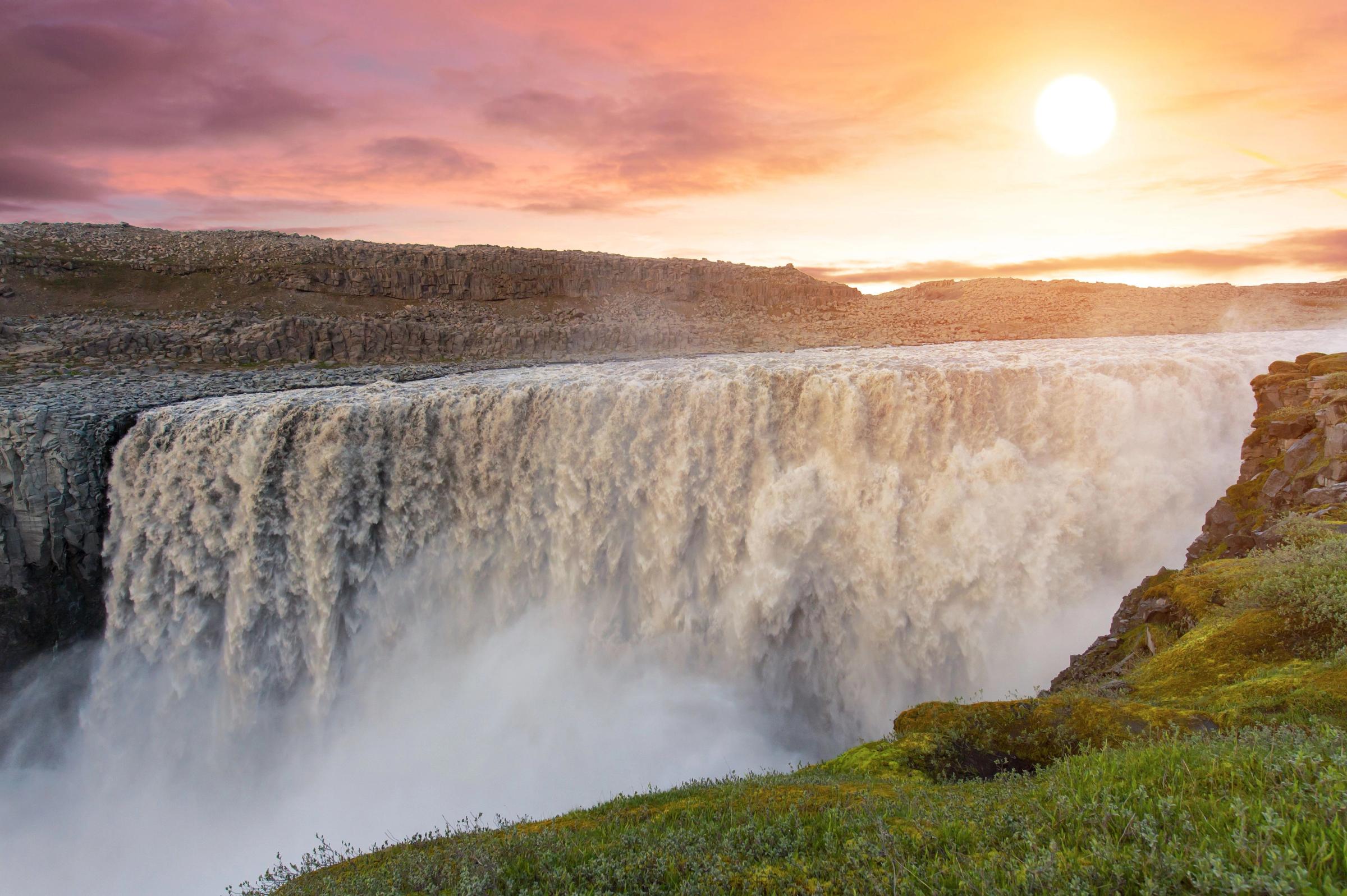 a waterfall in iceland at sunset with the sun shining through the clouds .