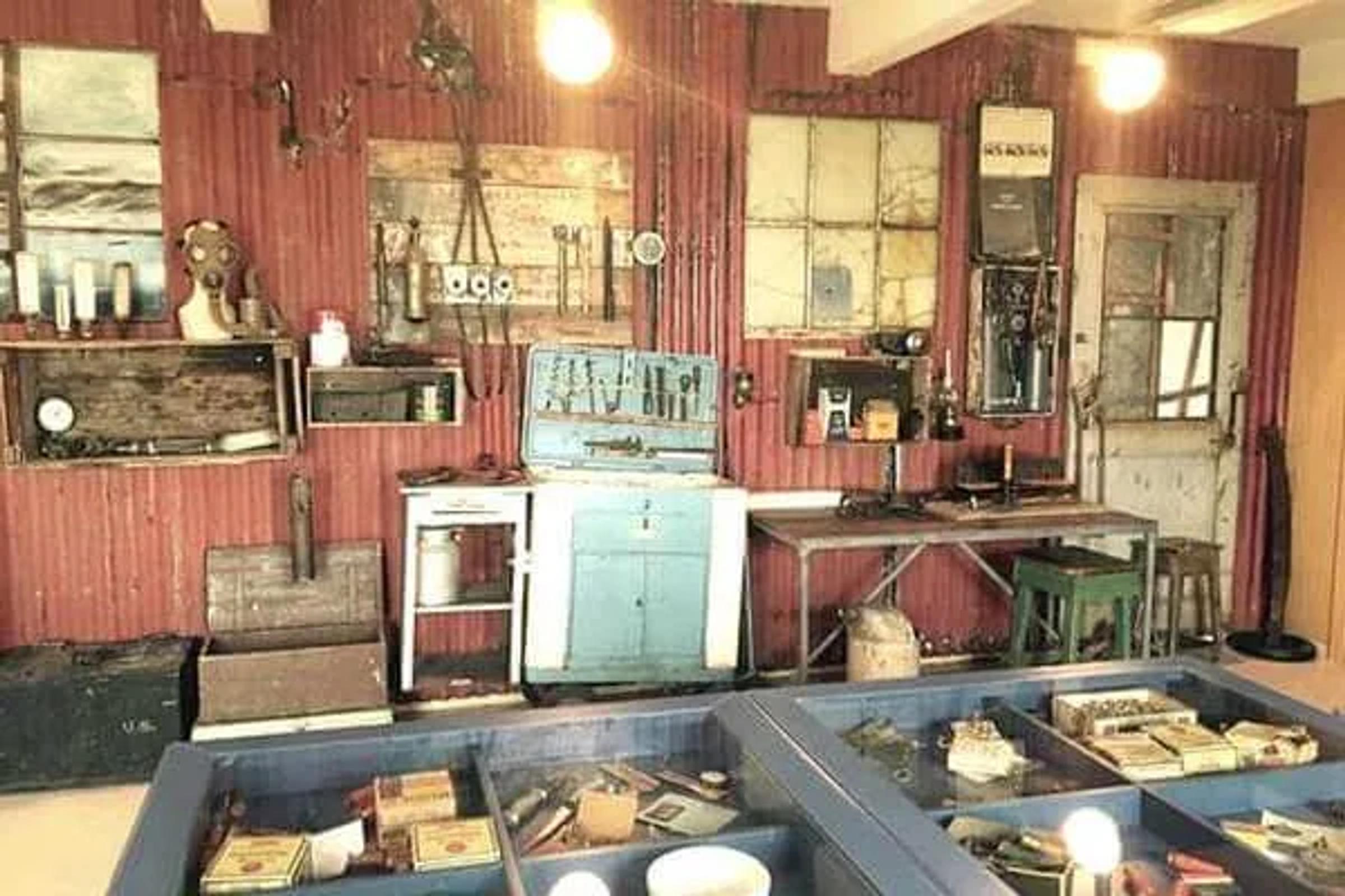 Un taller vintage o una exposición de museo con herramientas, maquinaria y objetos antiguos sobre paredes rojas de chapa ondulada y en vitrinas de cristal.