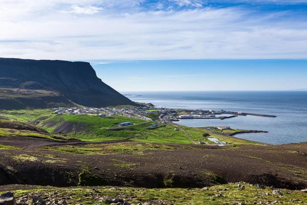 A small coastal town nestled at the base of a dark mountain, with a rocky foreground and blue sea.